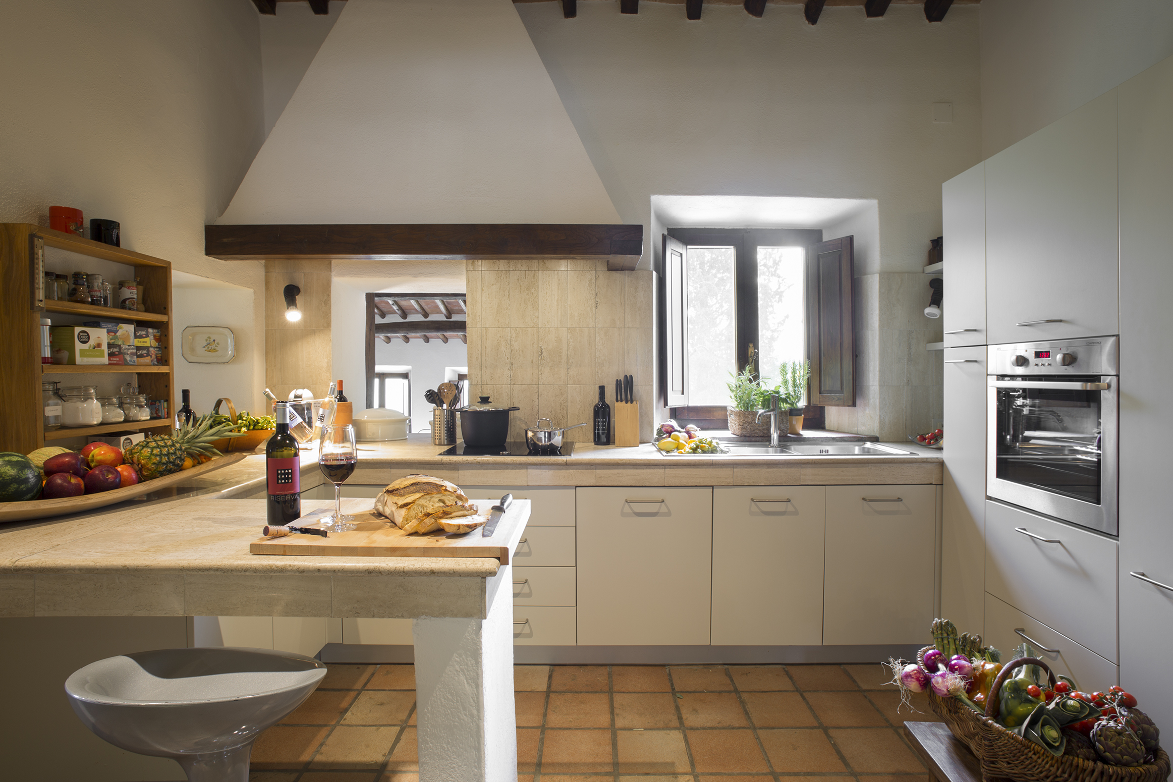 Una spaziosa cucina rustica con un tocco di stile toscano. A sinistra, una mensola piena di spezie, tè e erbe, accanto a una grande ciotola di frutta fresca, come mele, ananas e anguria. Sulla superficie della cucina c'è una bottiglia aperta di vino Brancaia e un bicchiere da vino accanto a un pane appena sfornato. Sullo sfondo, una finestra illumina la cucina, lasciando filtrare la luce del sole. La cucina è dotata di apparecchiature moderne ed emana un'atmosfera accogliente.