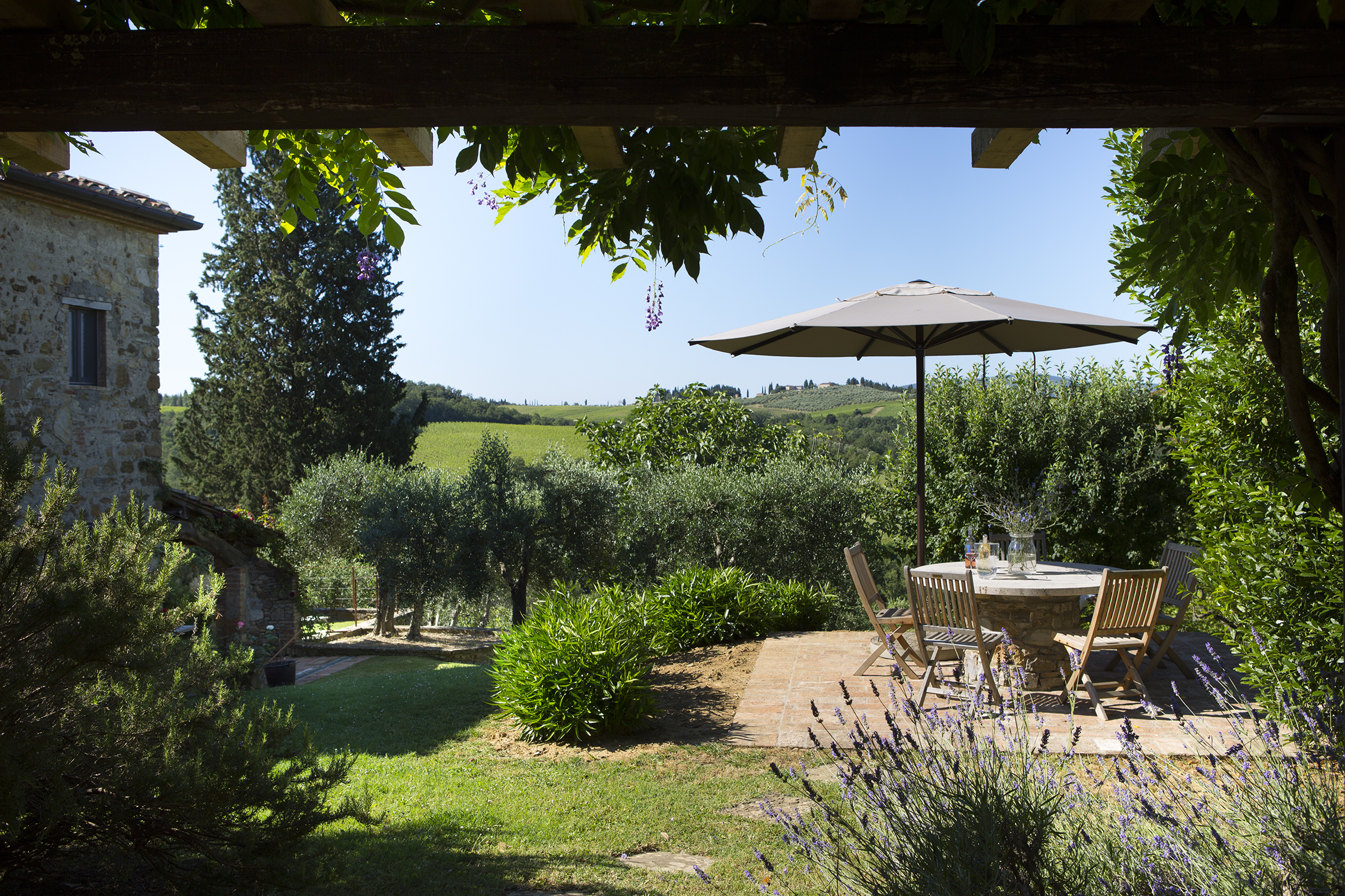 Una terrazza in pietra affascinante nel giardino, con vista sulle dolci colline del Chianti. Un tavolo rotondo in pietra con sedie in legno e un grande ombrellone offrono un luogo ombreggiato dove rilassarsi.<br><br> Lavanda e cespugli verdi incorniciano la scena, mentre sullo sfondo si vedono ulivi e cipressi, che sottolineano il tipico fascino toscano.