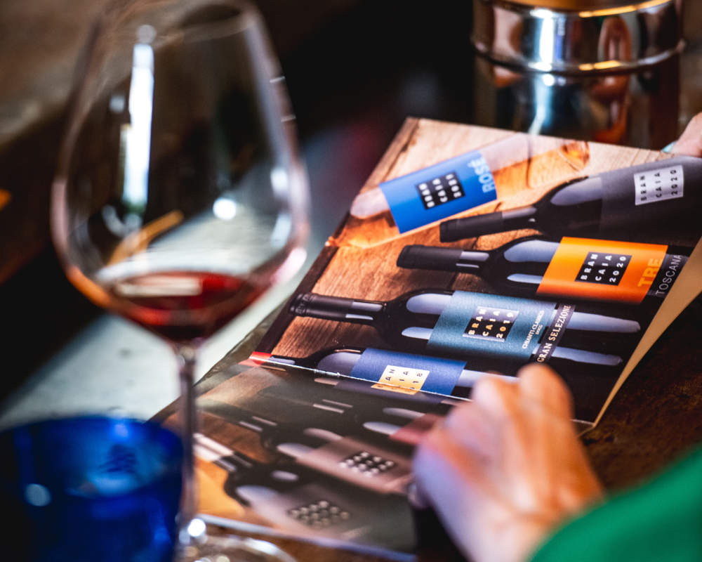 A hand points to the label of a Brancaia Chianti Classico bottle. The bottle features the typical black Brancaia label and a wine pourer on the opening. In the background, the person is wearing a Brancaia T-shirt. This scene symbolizes the expertise and attention to detail that is conveyed in the Brancaia wine tastings. Ideal for wine lovers who want to learn more about the production process of Chianti wines. Perfect for wine tours and guided visits at our winery in Tuscany.