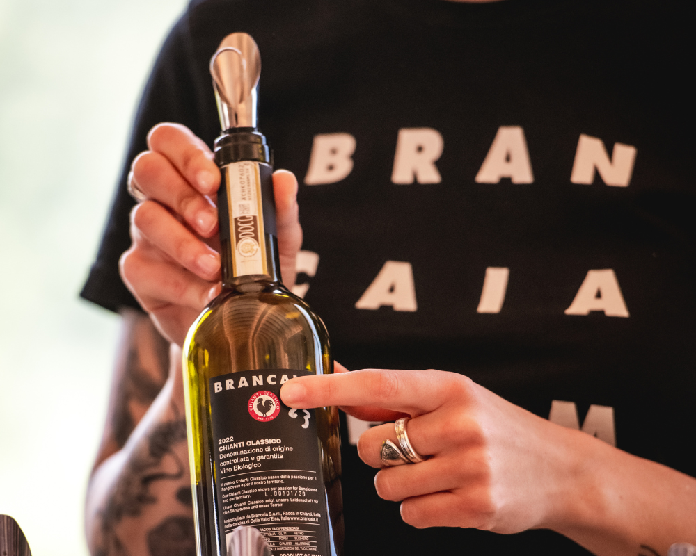 A hand points to the label of a Brancaia Chianti Classico bottle. The bottle features the typical black Brancaia label and a wine pourer on the opening. In the background, the person is wearing a Brancaia T-shirt. This scene symbolizes the expertise and attention to detail that is conveyed in the Brancaia wine tastings. Ideal for wine lovers who want to learn more about the production process of Chianti wines. Perfect for wine tours and guided visits at our winery in Tuscany.
