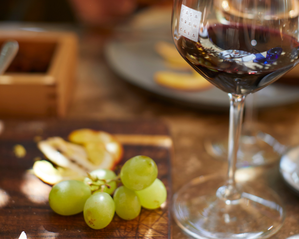 A glass of red wine with the Brancaia logo on a table, next to it a wooden board with green grapes and thinly sliced cheese. A cozy atmosphere with warm light.