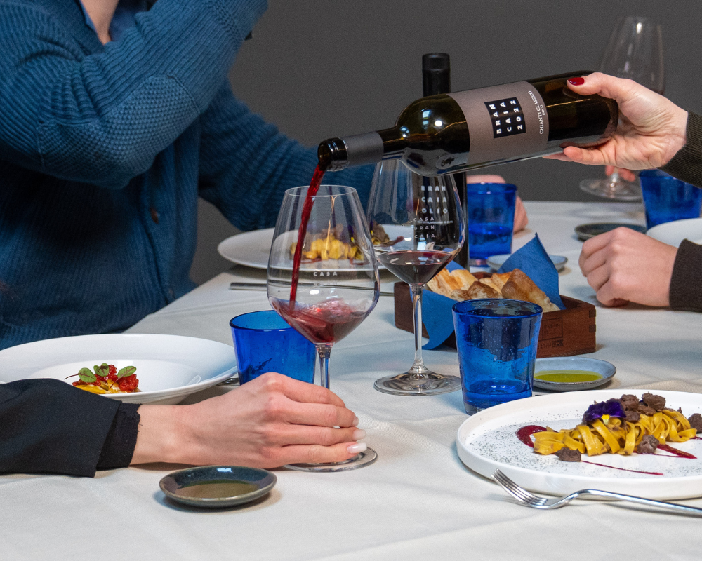 A bottle of wine with the Brancaia label is poured into a glass while several people are seated at a table set with plates, bread and blue glasses. An elegant and convivial atmosphere.