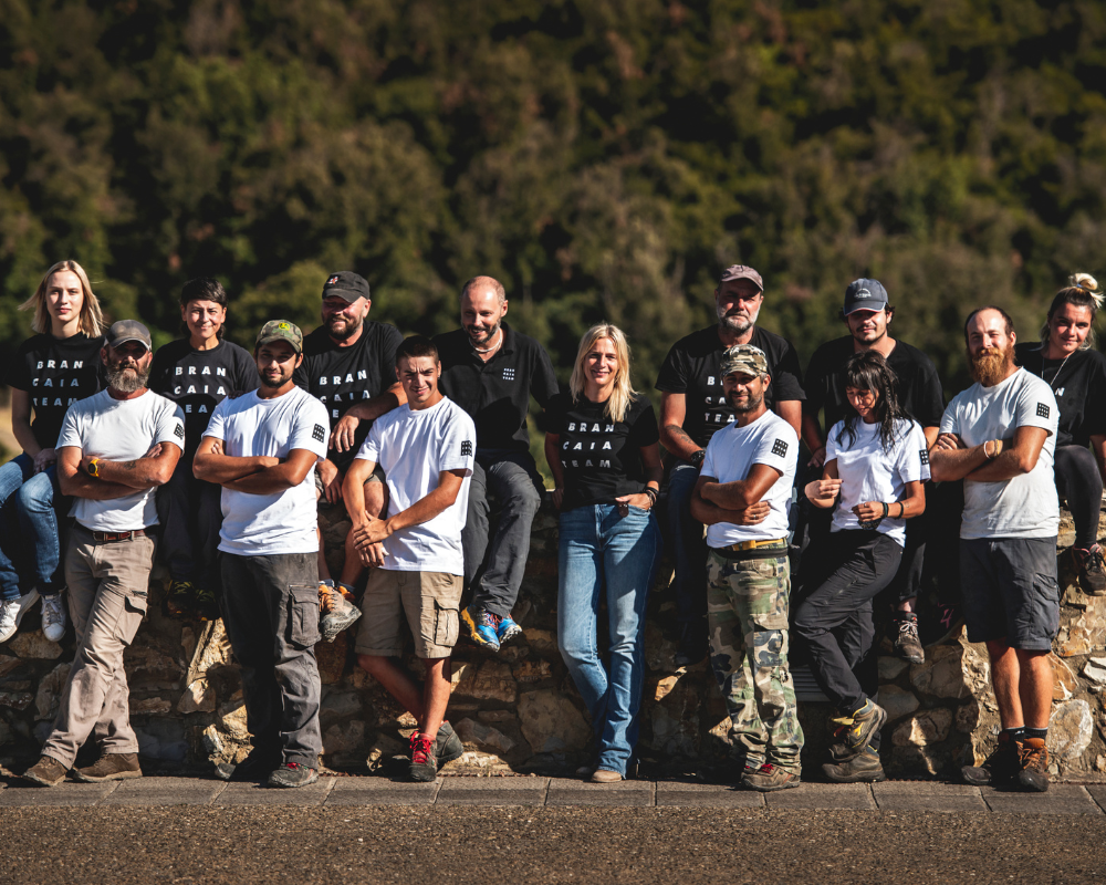 Das Brancaia-Team steht auf einer Steinmauer im Freien vor einer grünen, bewaldeten Landschaft. Die Teammitglieder tragen schwarze und weiße T-Shirts mit dem Brancaia-Logo und lächeln stolz in die Kamera. Sie zeigen eine starke Gemeinschaft und Engagement für die Arbeit in der Weinproduktion.
